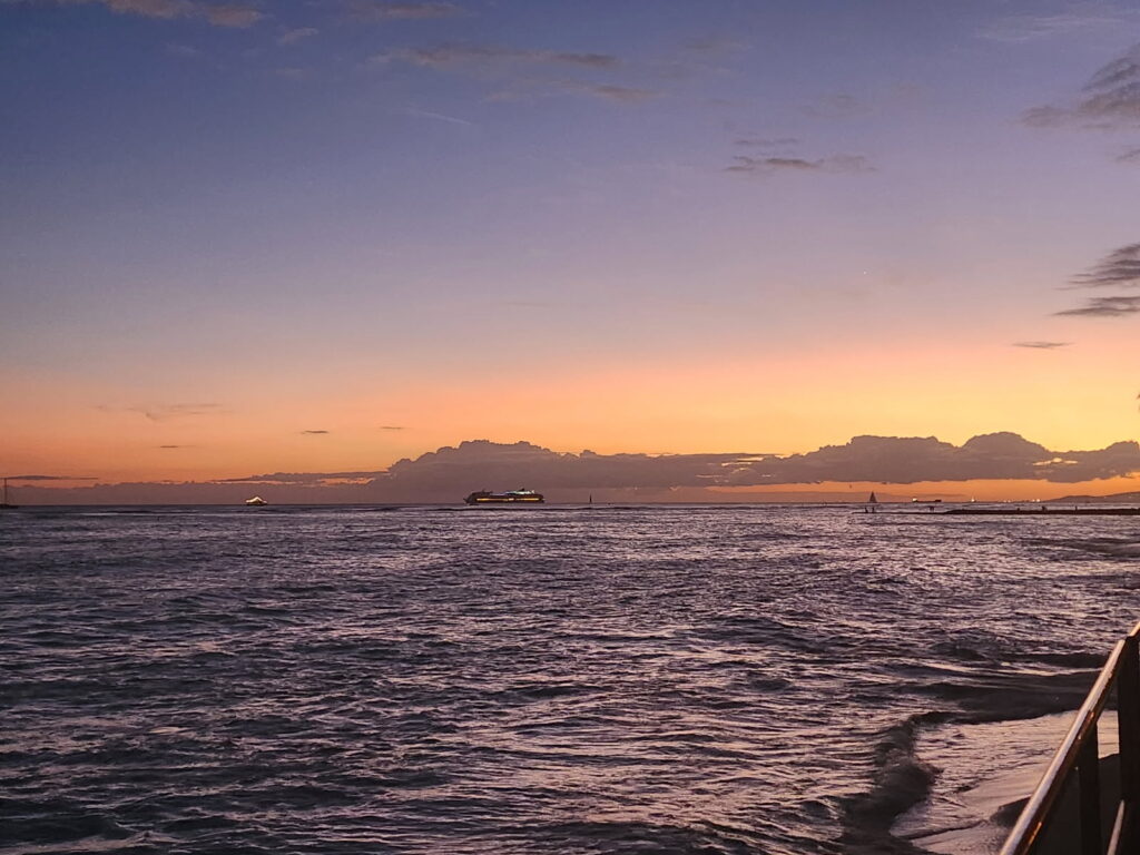 The sun sets over the ocean, casting warm hues across the water, as seen from the deck of a boat.